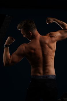 Shirtless man flexing muscles against a dark background, showcasing physique.