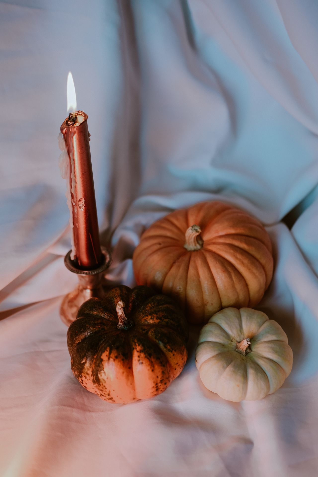 Lit red taper candle in a brass holder beside orange, dark, and white pumpkins on draped fabric