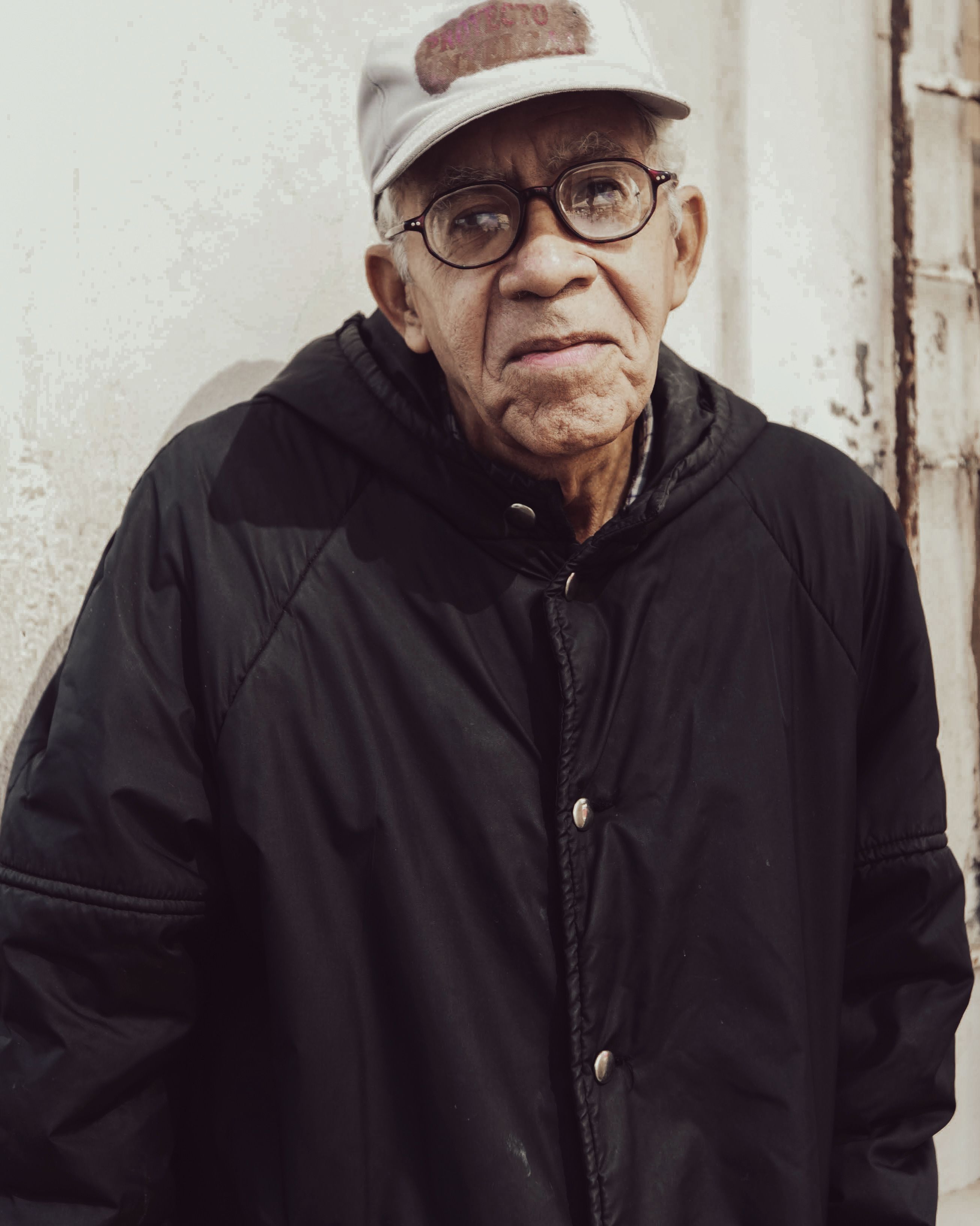 Elderly man wearing glasses and a baseball hat, standing outdoors in a black jacket.
