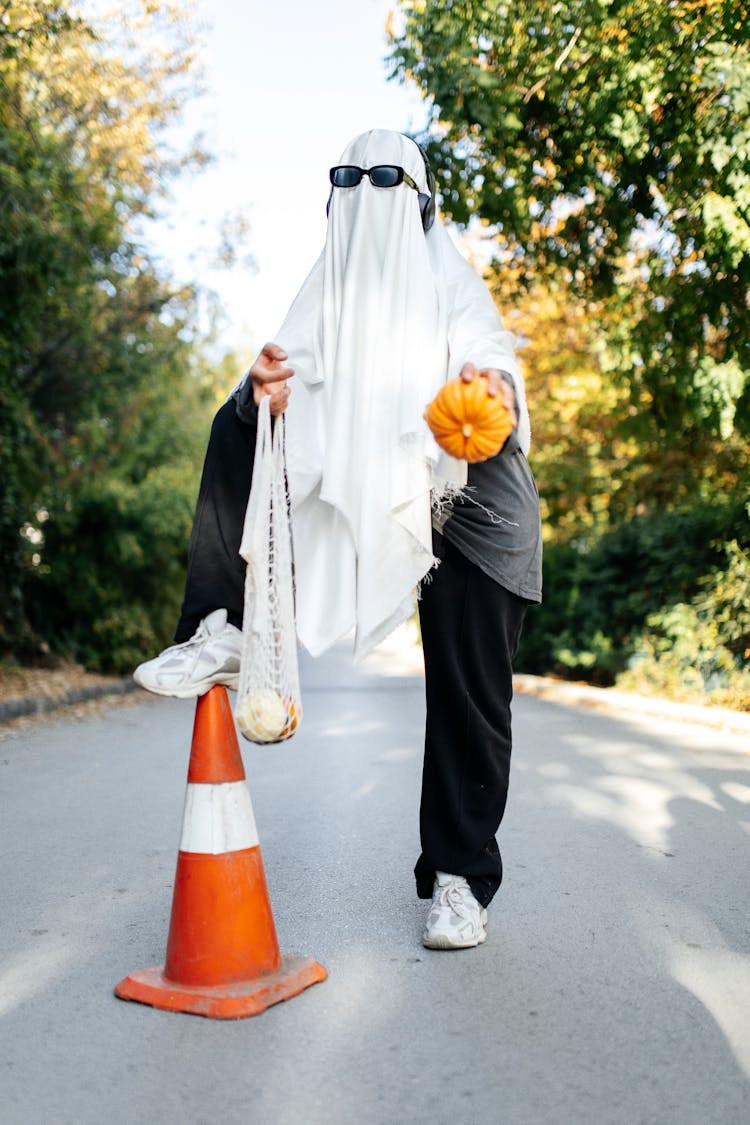 Man In A Ghost Costume Holding Small Pumpkin And Shopping Net Standing On A Traffic Cone