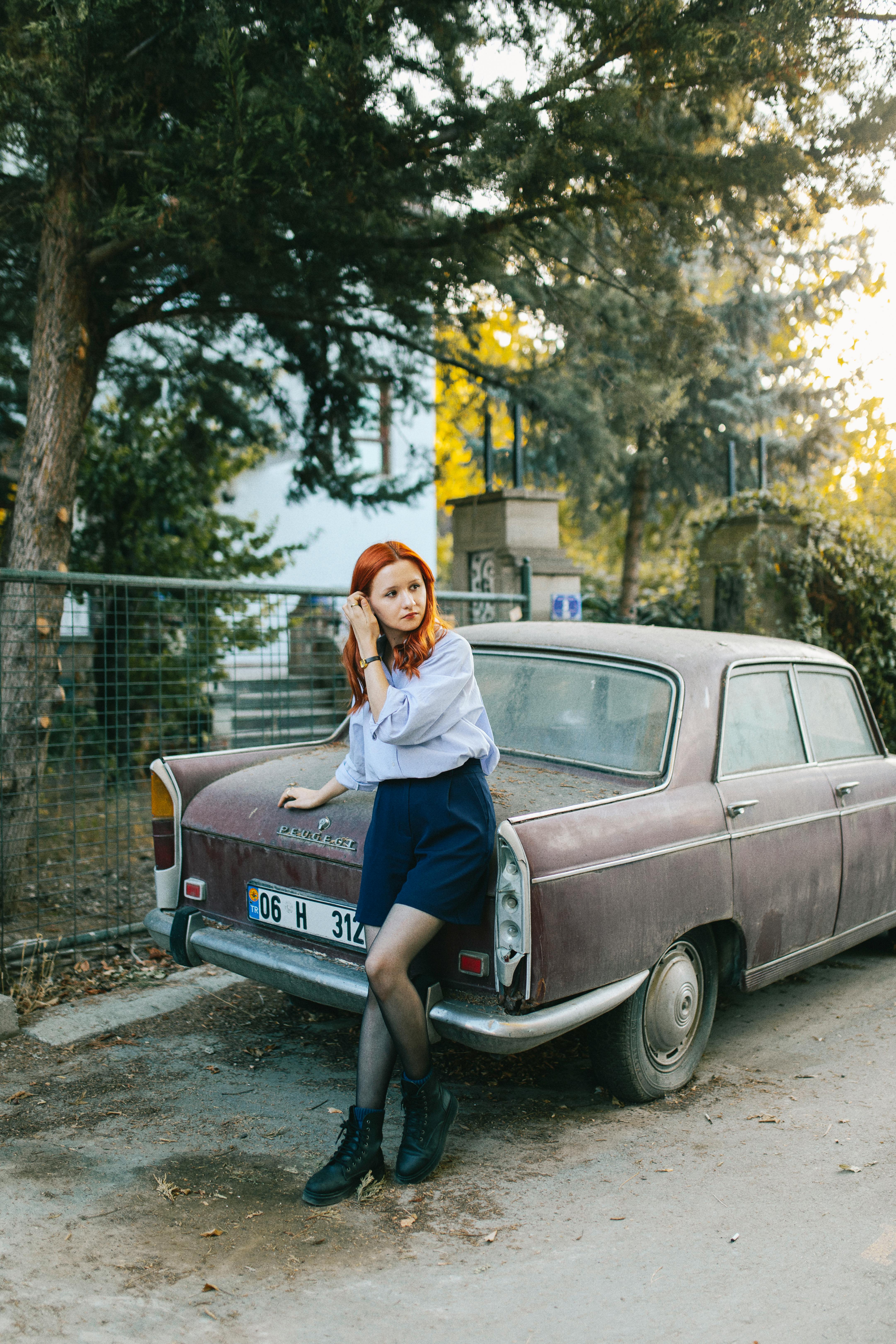 Redhead woman in fashion outfit posing with a vintage burgundy car outdoors, exuding retro charm.