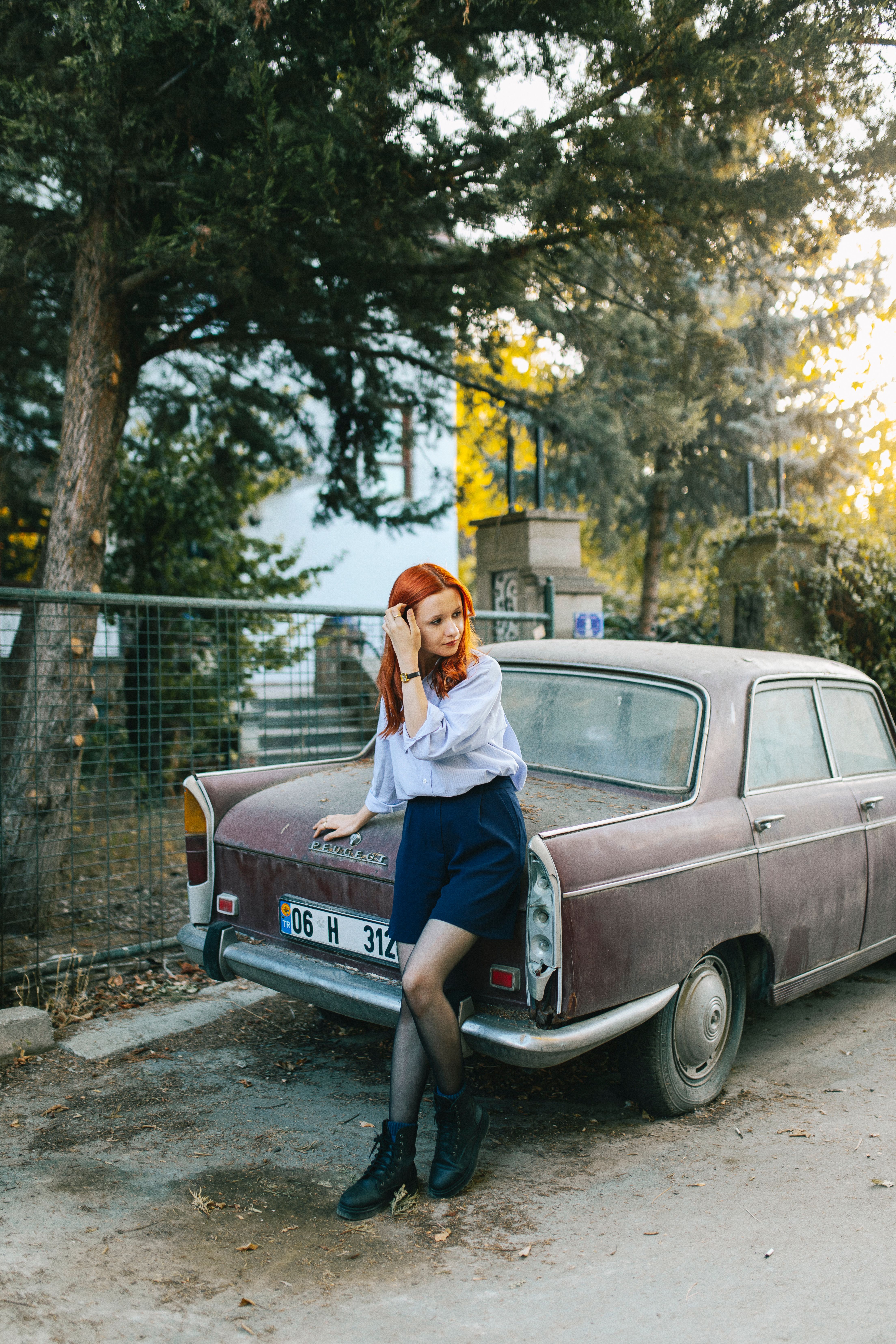 Red-haired woman in stylish attire posing by a retro car in a natural setting.