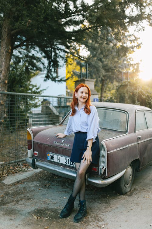 Smiling Young Woman in a Purple Blouse and Navy Blue Mini Skirt Leaning on Old Abandoned Car