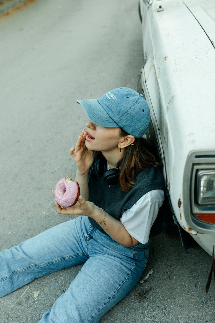 Young Woman Eating A Glazed Donut Sitting On The Asphalt Leaning Against The Wheel Of A Car