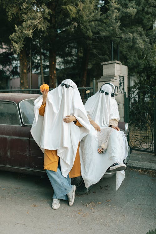 Two People Wearing Simple Ghost Costumes Sitting on a Parked Car with Pumpkins in Hands