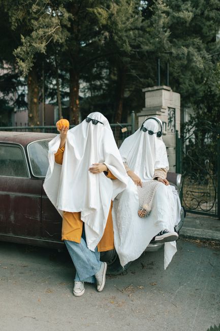 Two people in ghost costumes with pumpkins lean against a vintage car in a humorous Halloween setting.