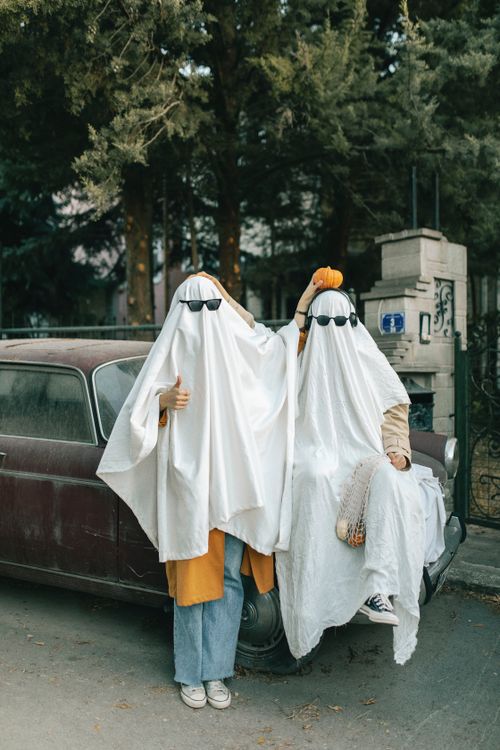 Free Two people dressed as ghosts with sunglasses in front of an old car, celebrating Halloween. Stock Photo
