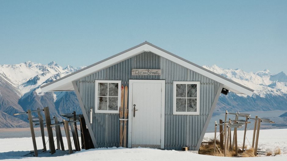 A solitary alpine cabin in a snowy landscape with skis leaned against it.