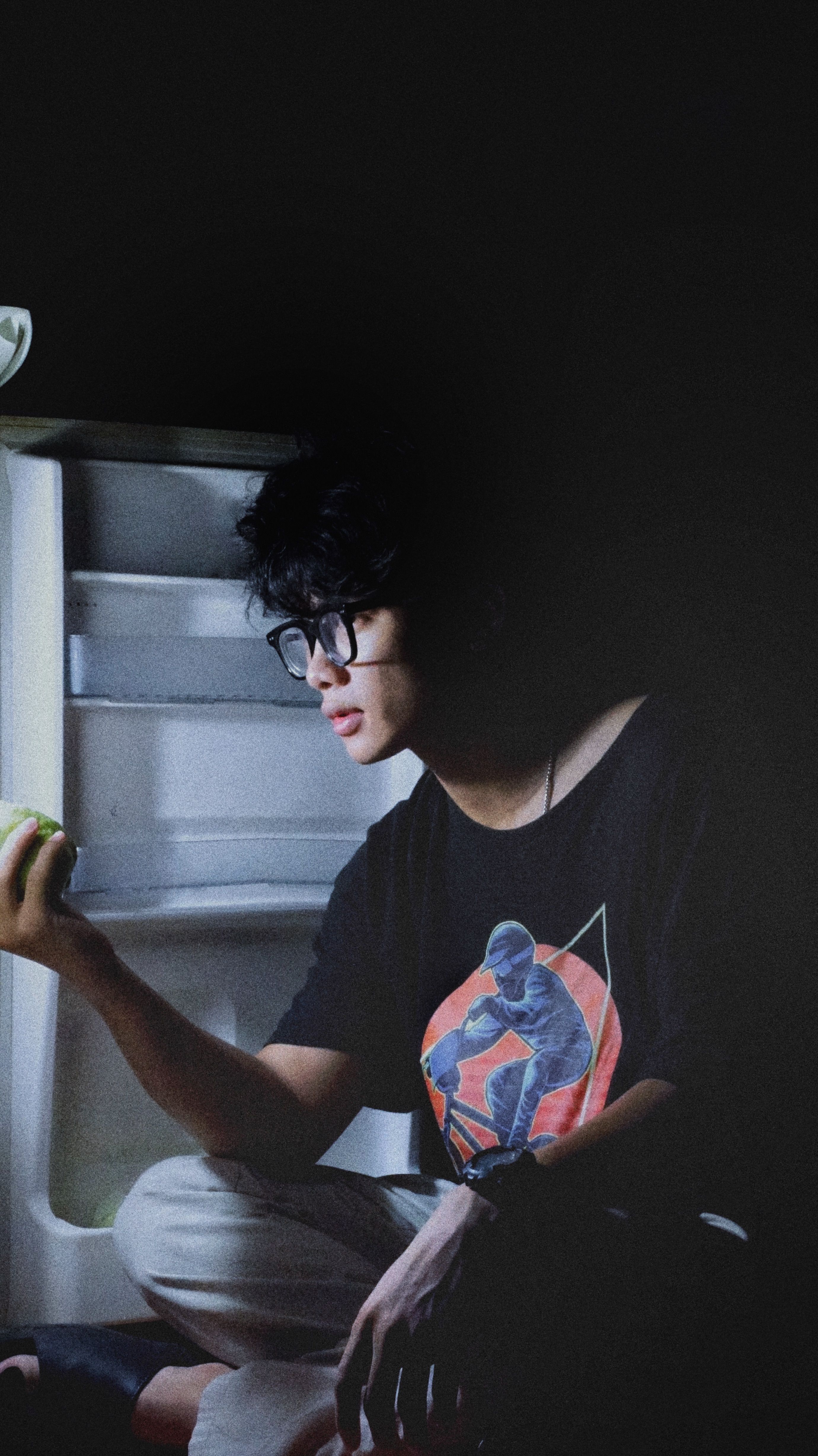 A young man in eyeglasses sits illuminated by an open refrigerator light, holding an apple.