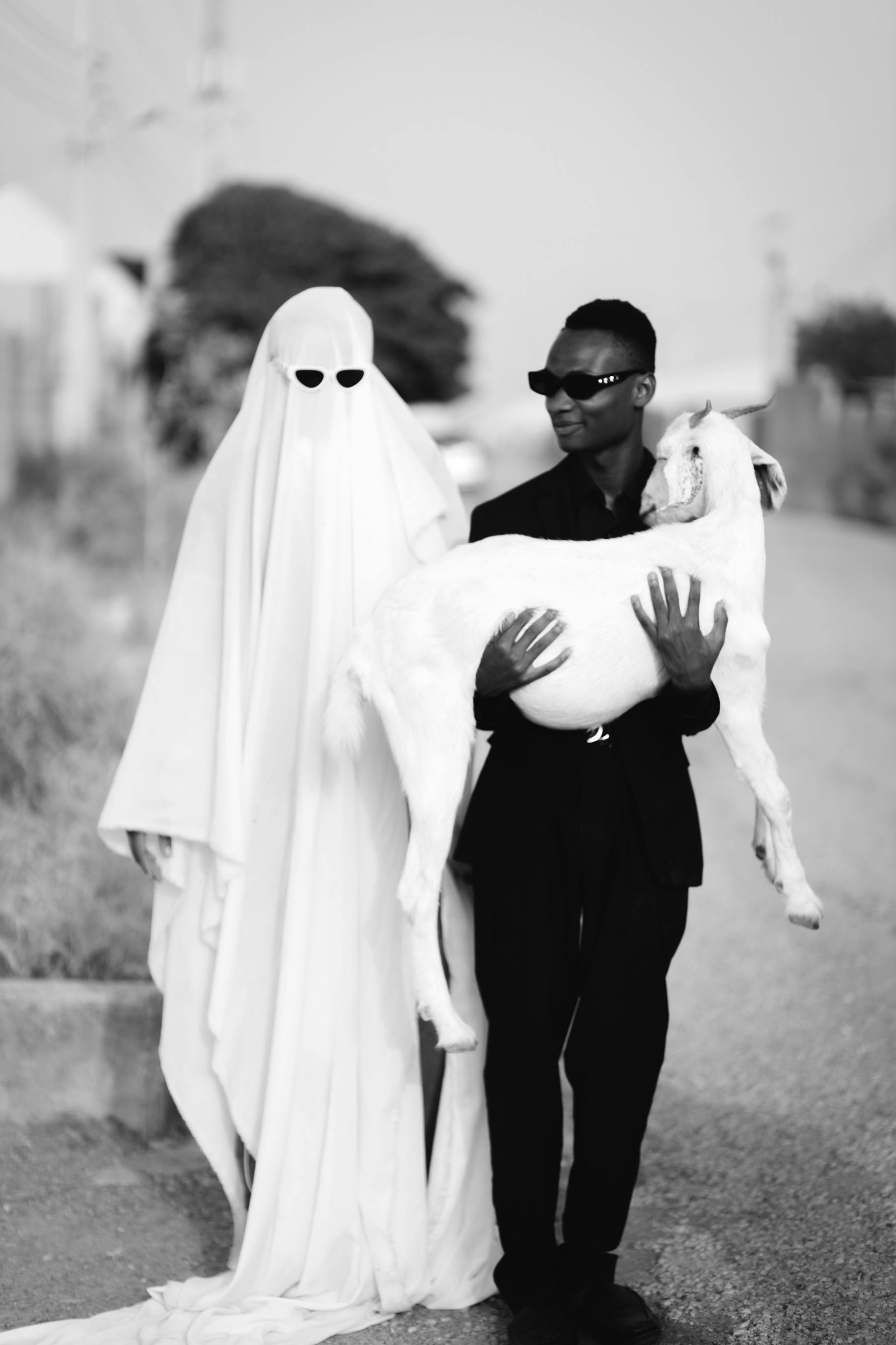 Person Covered in a White Sheet Standing behind a Young Man Carrying a Goat · Free Stock Photo