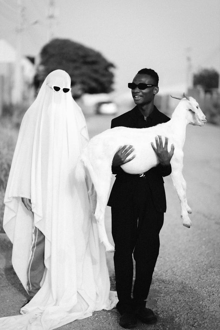 Man In Sunglasses Holding Goat Doll And Standing With Ghost On Road In Village