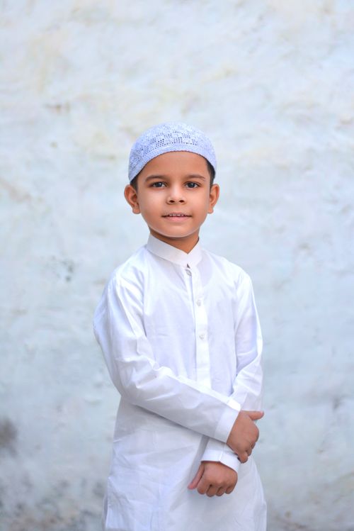 Free A portrait of a boy wearing traditional white Islamic clothing and cap, standing indoors. Stock Photo