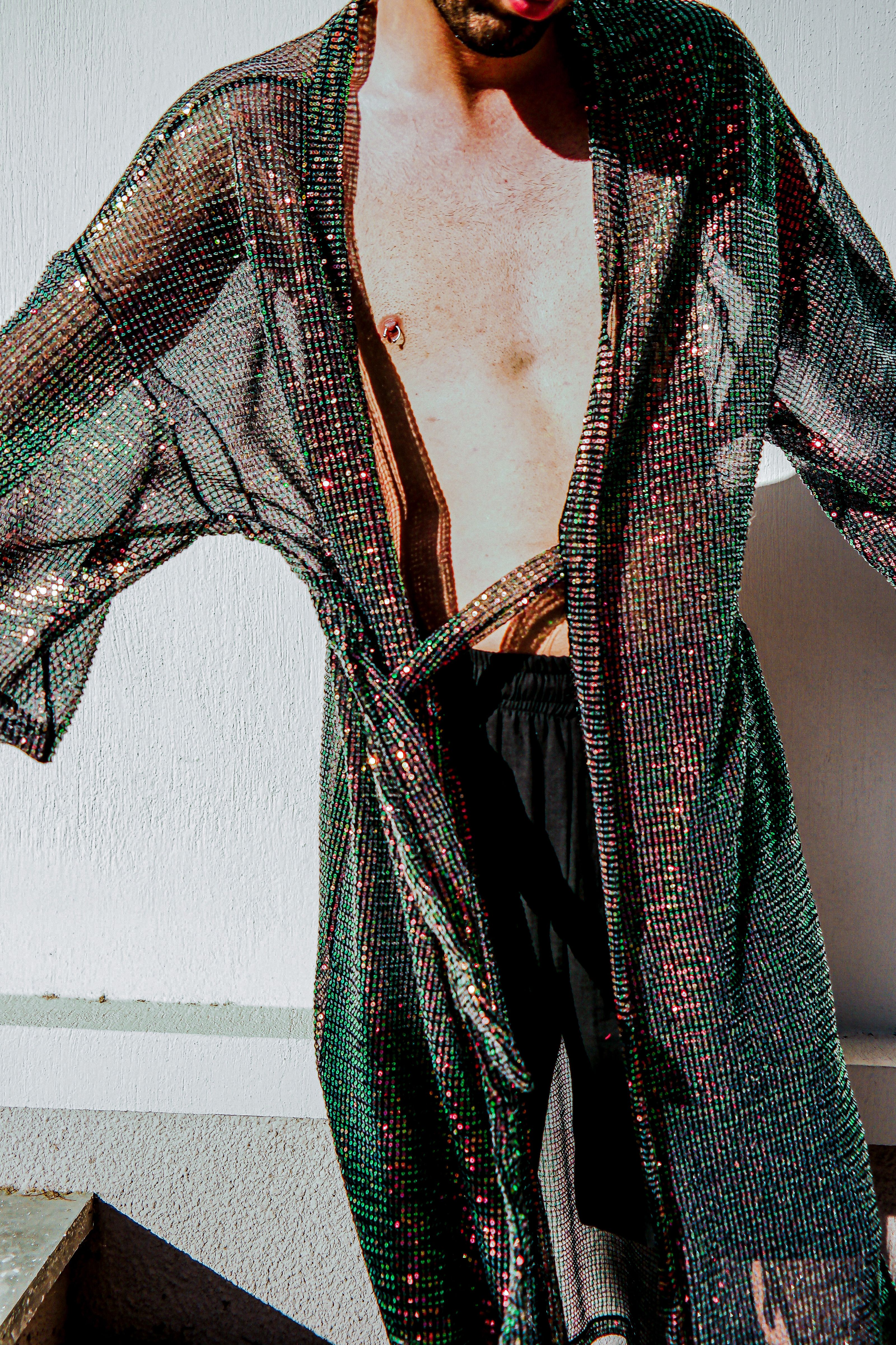 Um homem fashion usando um robe brilhante e transparente posa sem camisa dentro de casa.