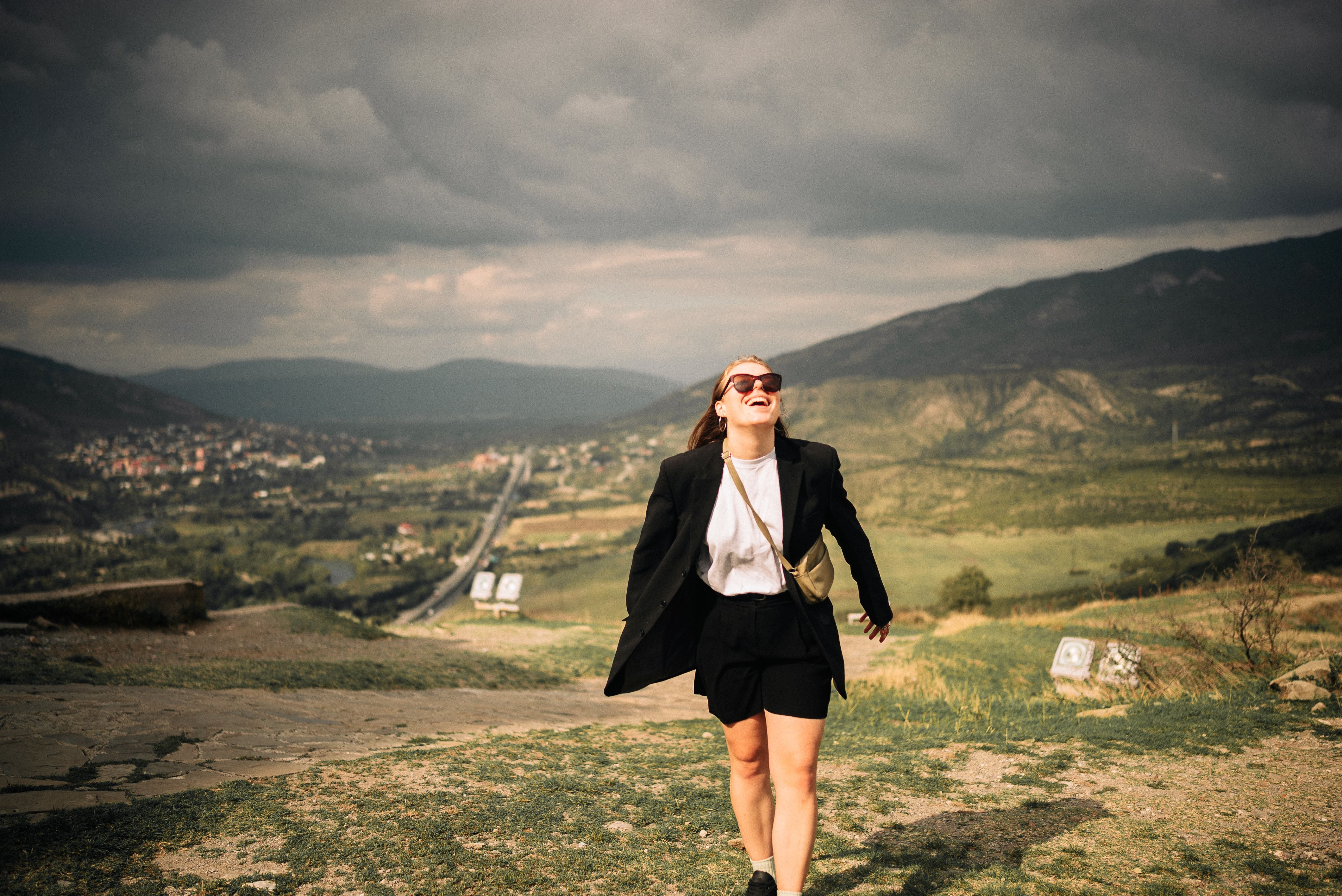 Fashionable woman with black jacket and sunglasses walking in a scenic countryside landscape, smiling and enjoying nature.