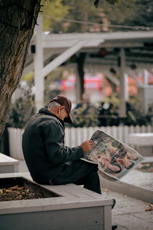 Free Senior man reading a newspaper on a park bench, captured during the day with a city backdrop. Stock Photo