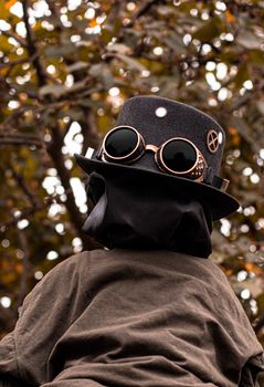 Intriguing steampunk outfit featuring goggles, hat, and dark cloak set against a blurred fall backdrop.