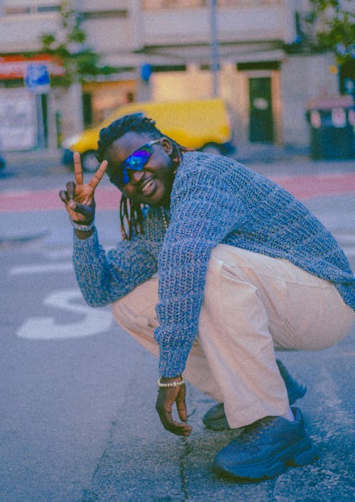 Musician in a Knitted Blue Sweater and Sunglasses Showing V-sign Crouching on a Street