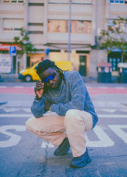 Musician in a Knitted Blue Sweater and Sunglasses Crouching at a Stop Sing on the Street