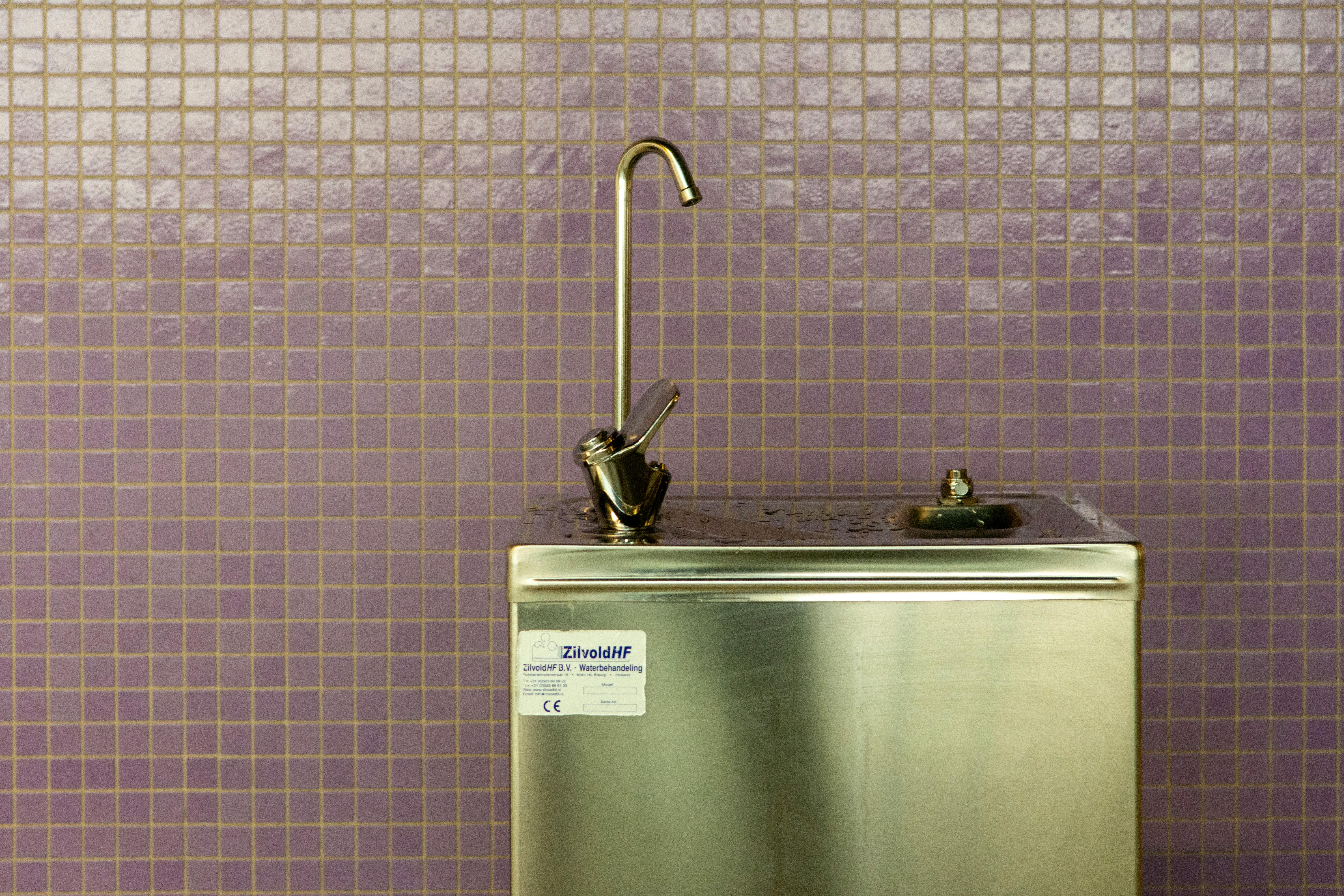 A stainless steel drinking fountain set against a purple mosaic tiled wall, urban environment.