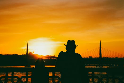 Silhouette of a man in a fedora watching a vibrant orange sunrise over a suspension bridge.