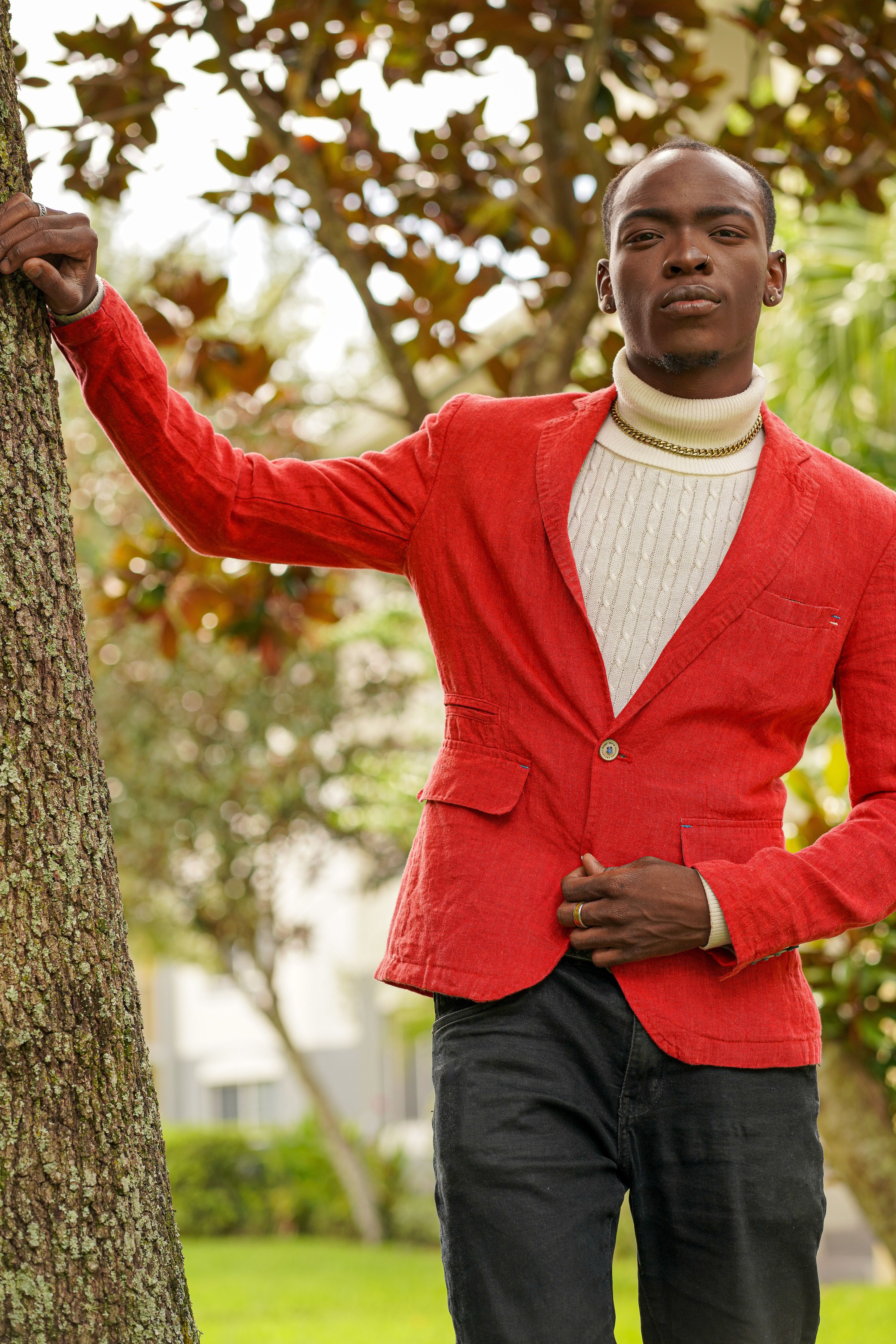 Young man in a red jacket posing by a tree in a park, showcasing modern fashion.