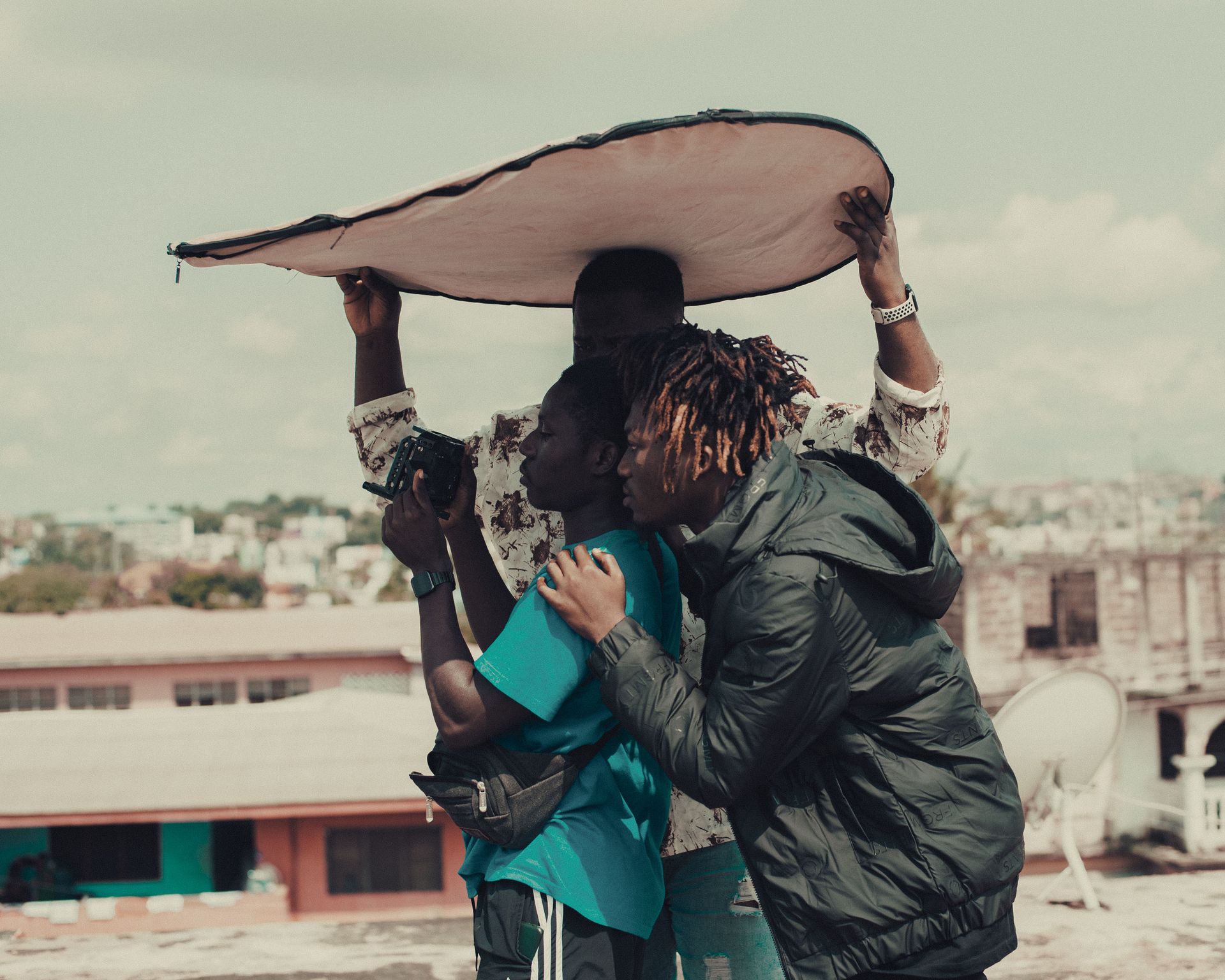 Three crew members adjust camera settings under a reflector on an urban rooftop.