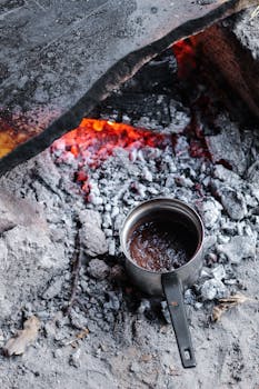 Metal pot brewing coffee over an outdoor campfire, surrounded by glowing embers and charcoal.