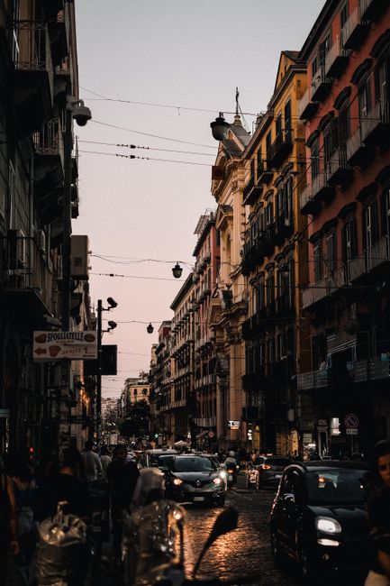 Cars navigate a busy, narrow street lined with historic buildings in a European city at twilight.