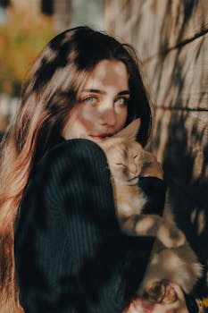 Charming portrait of a young woman with long hair, holding a cat, bathed in warm sunlight.