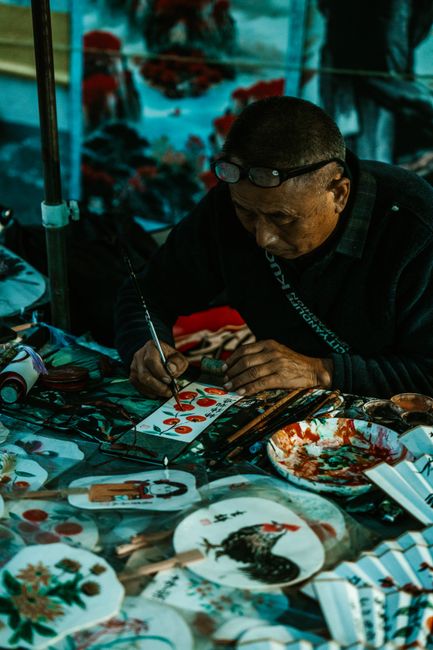 An artist focused on detailed painting at a cluttered table, showcasing traditional art forms.