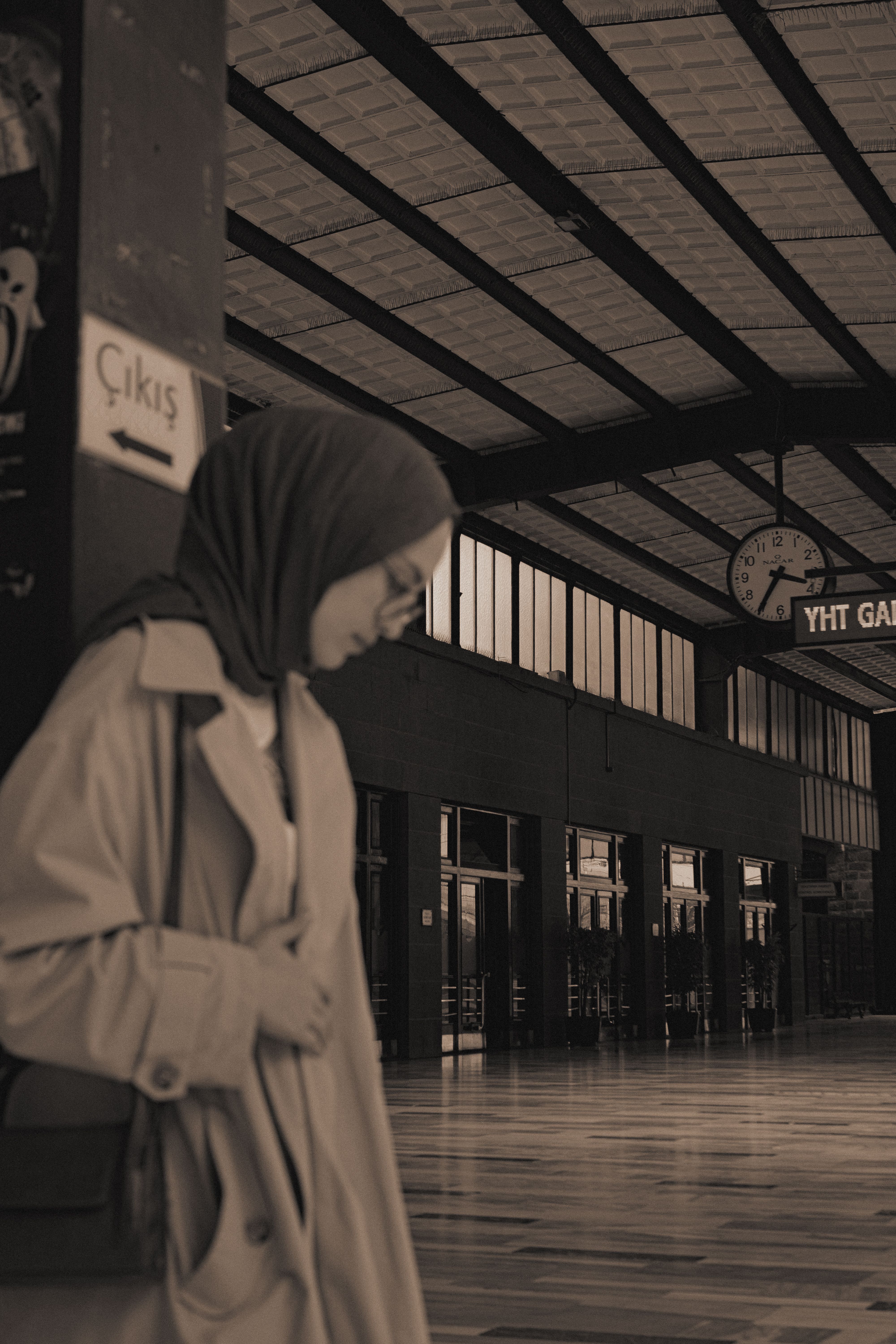 A woman in a headscarf stands in a sepia-toned railway station hallway.