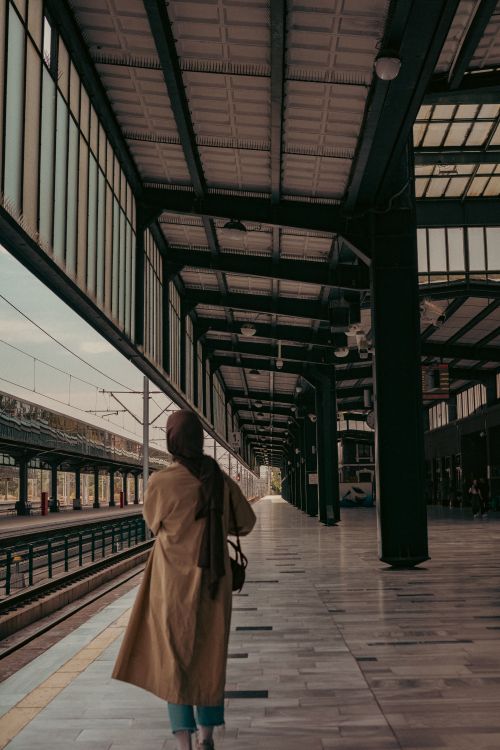 Free A woman in a hijab and trench coat walking along a modern train station platform. Stock Photo