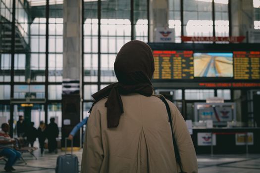 Back view of a woman in hijab observing the train schedule in a modern station.