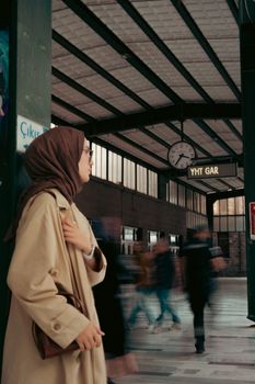 A woman in hijab stands at a busy railway station with blurred pedestrians in motion.