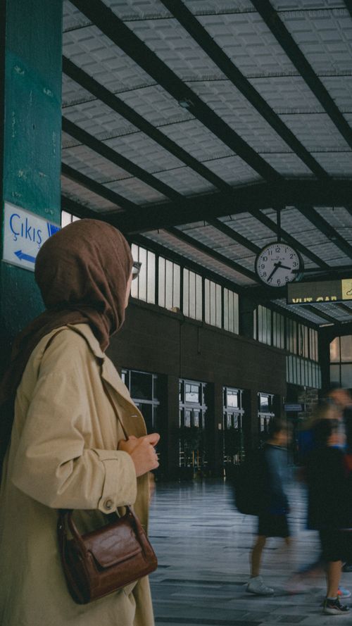 Free A woman wearing a hijab stands on a bustling train station platform, observing the blurred motion of passing pedestrians. Stock Photo