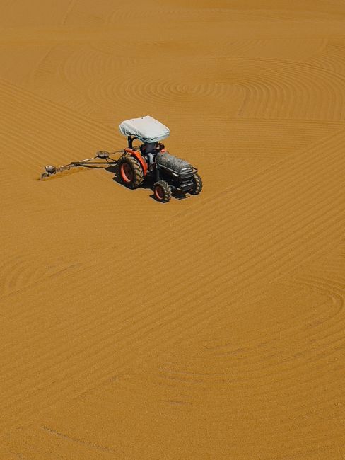 A tractor plows through a vast sandy landscape, creating intricate patterns from an aerial perspective.