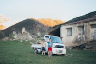 在一座以山为背景的乡村建筑旁，两个孩子坐在一辆白色卡车旁边