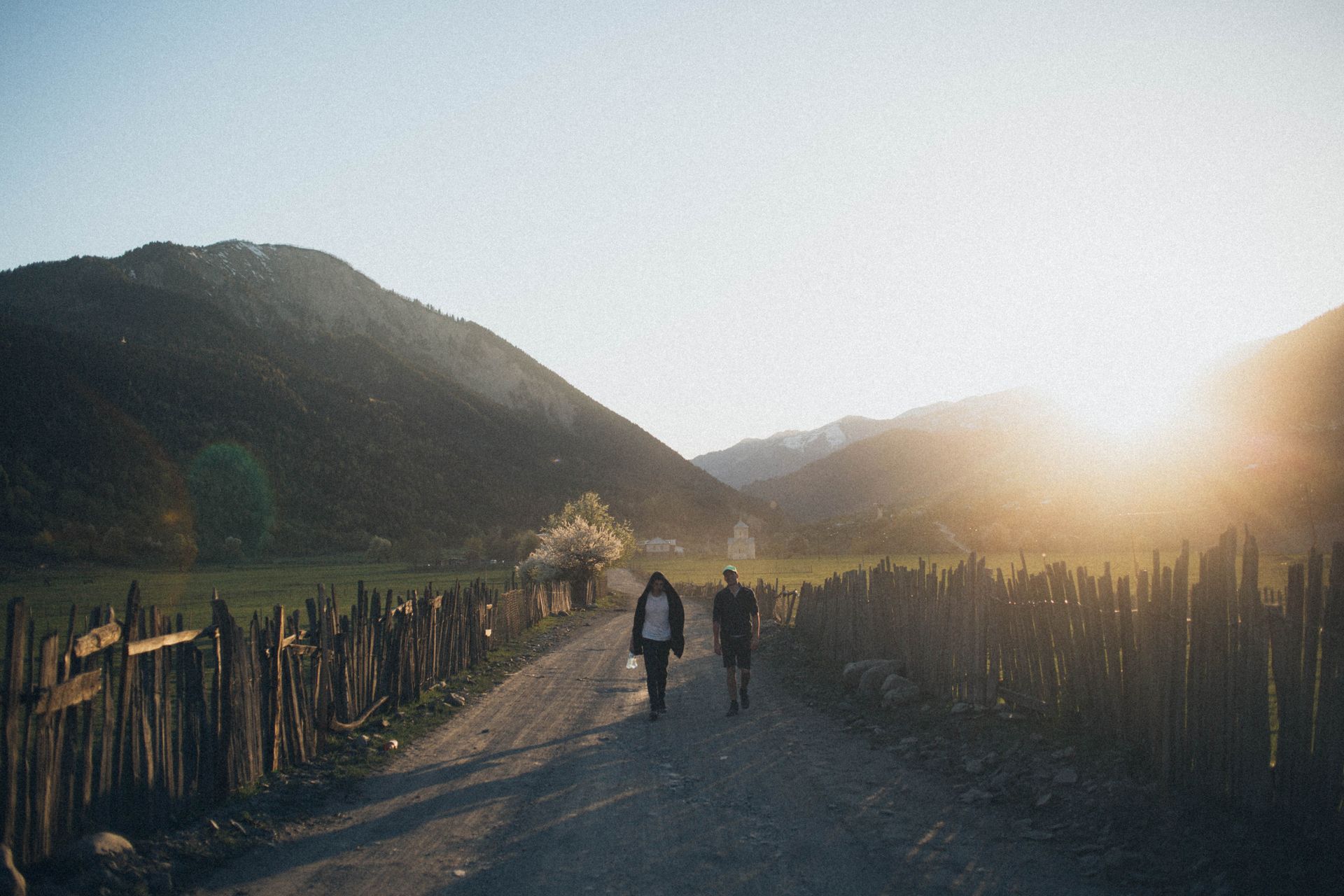 Two people walking on a rural dirt road at sunset, surrounded by mountains and wooden fences.