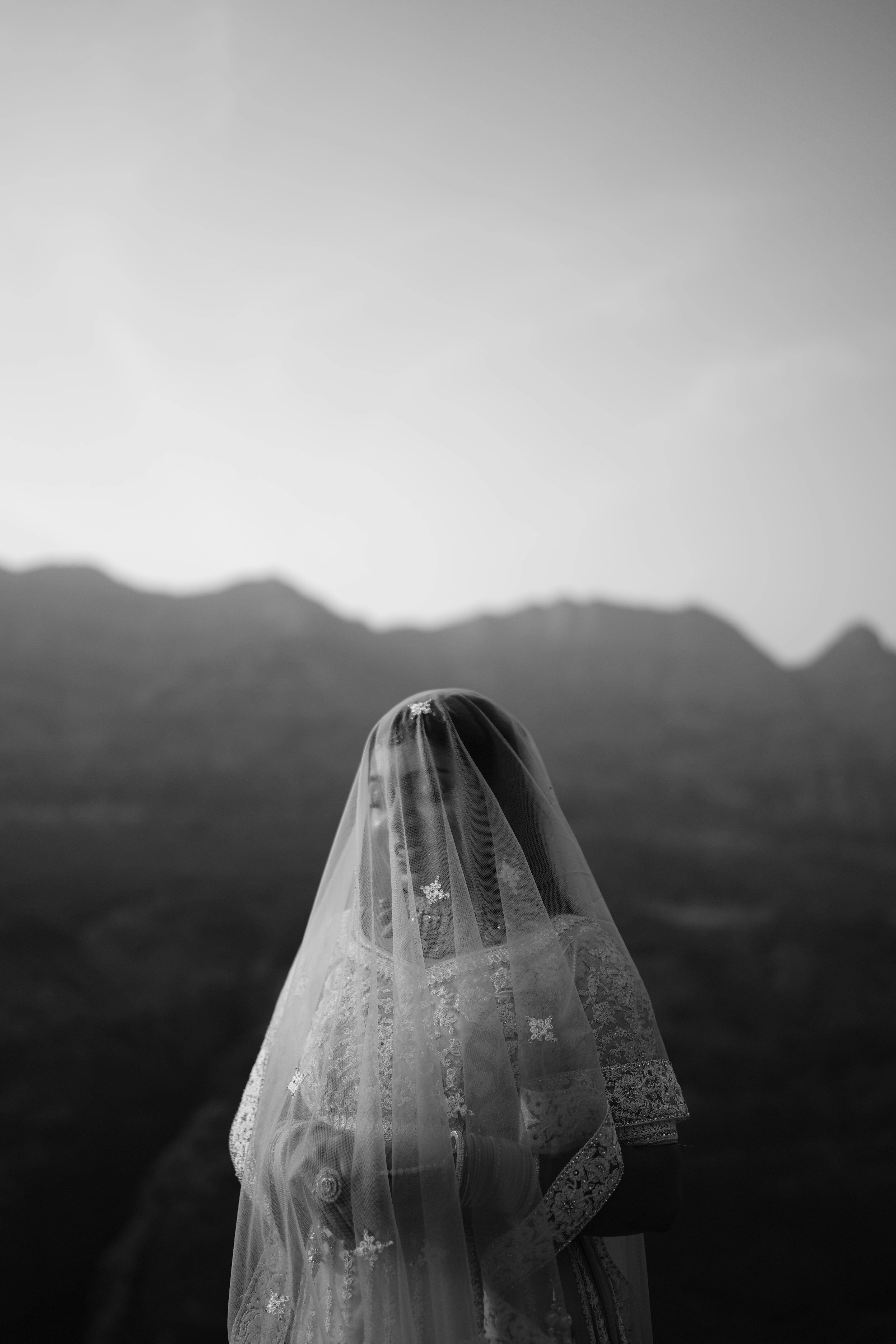 Black and white portrait of a bride with veil and lace, set against mountains.