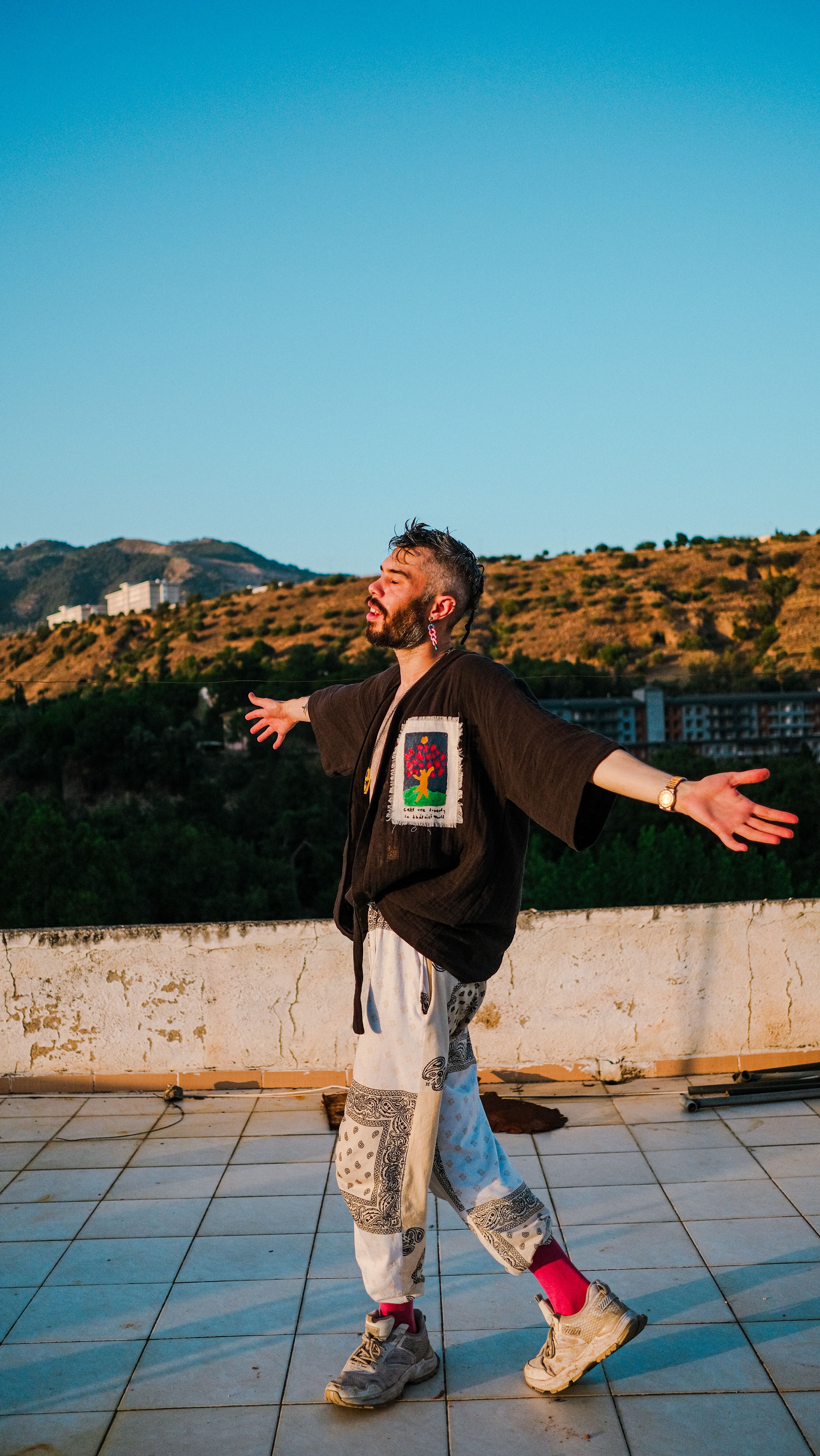 A young man in stylish attire joyfully poses with arms open wide outdoors under sunny skies.