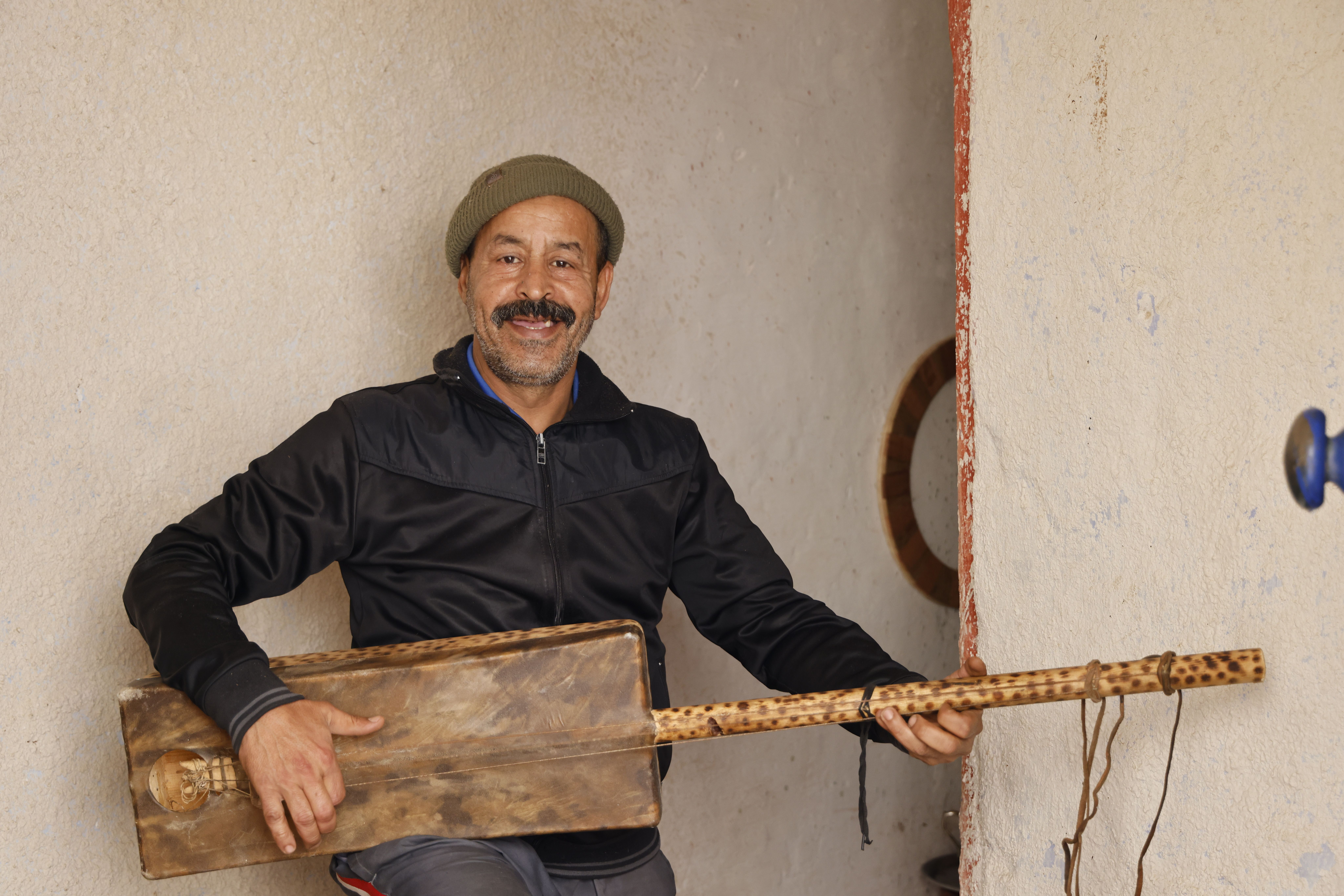 A smiling man in a hat and black jacket holds a handmade musical instrument.