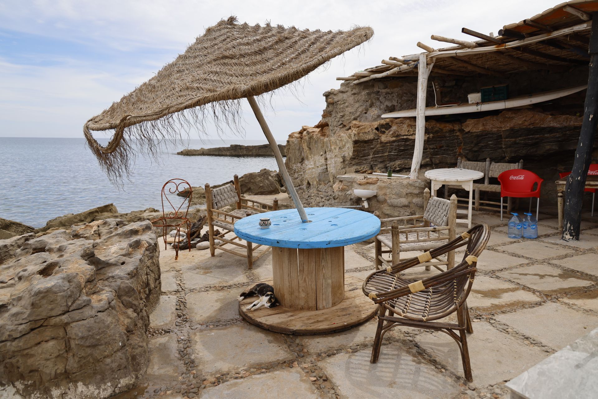 A panoramic view of a rustic seaside café with eclectic furniture and a natural umbrella.