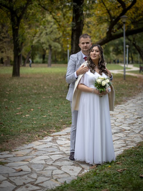 Newlyweds Standing Together in Alley in Park