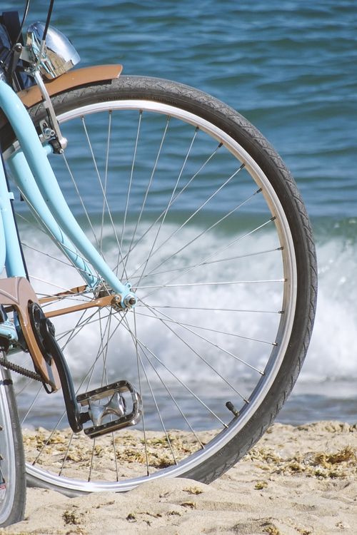 Free Close-up of a bicycle wheel on a sandy beach with ocean waves in the background. Stock Photo