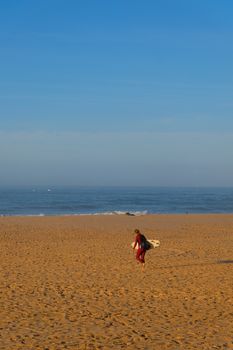 A surfer carrying a surfboard walks along Carcavelos Beach in Portugal during a sunny day.
