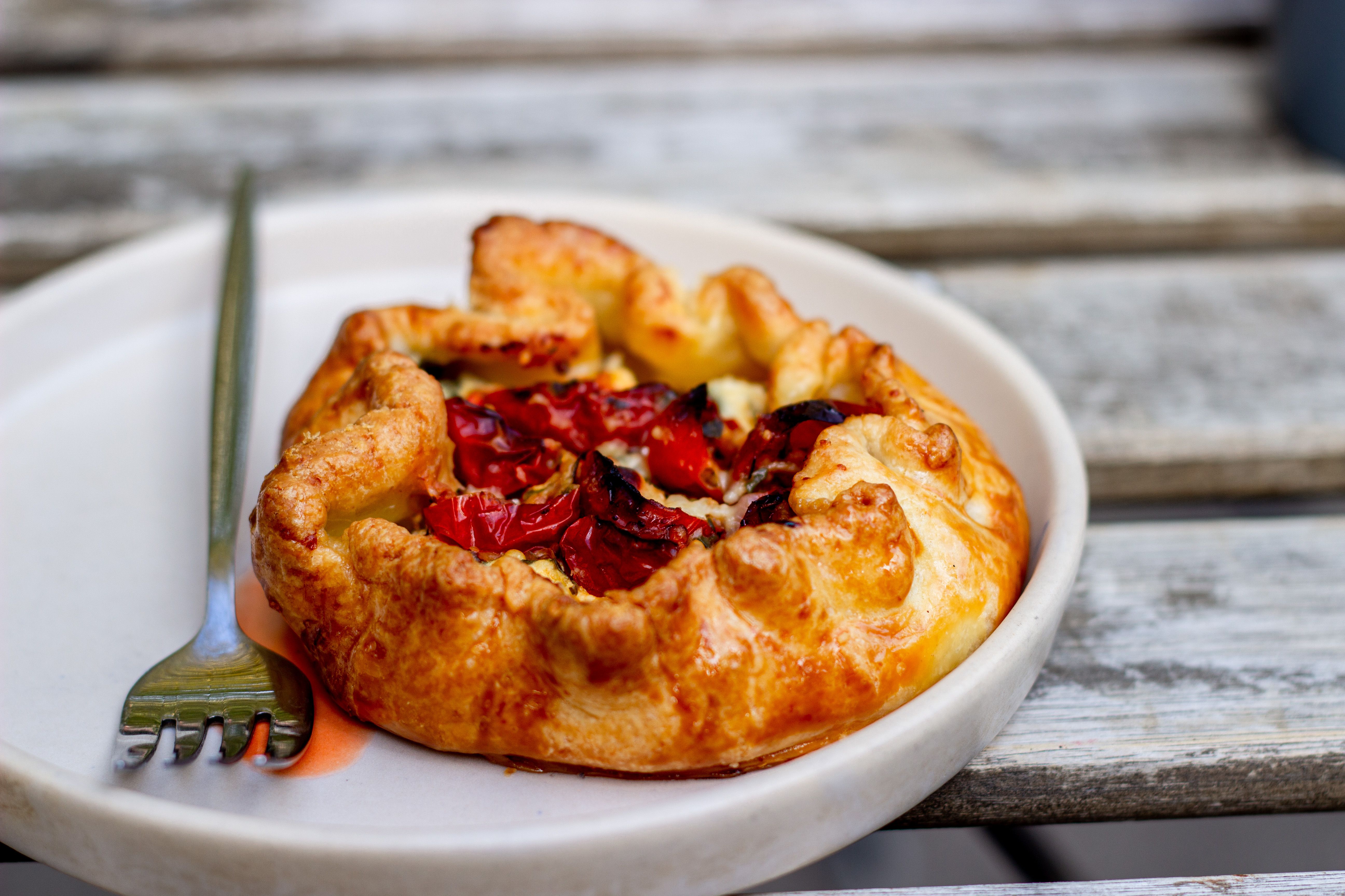 A golden, puffed pastry tart topped with overlapping tomato slices, crumbled goat cheese, and fresh thyme, sliced into squares on a wooden board.