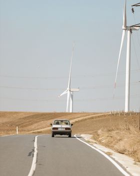 A classic car drives down a rural road lined with wind turbines, depicting sustainable energy in a countryside setting.
