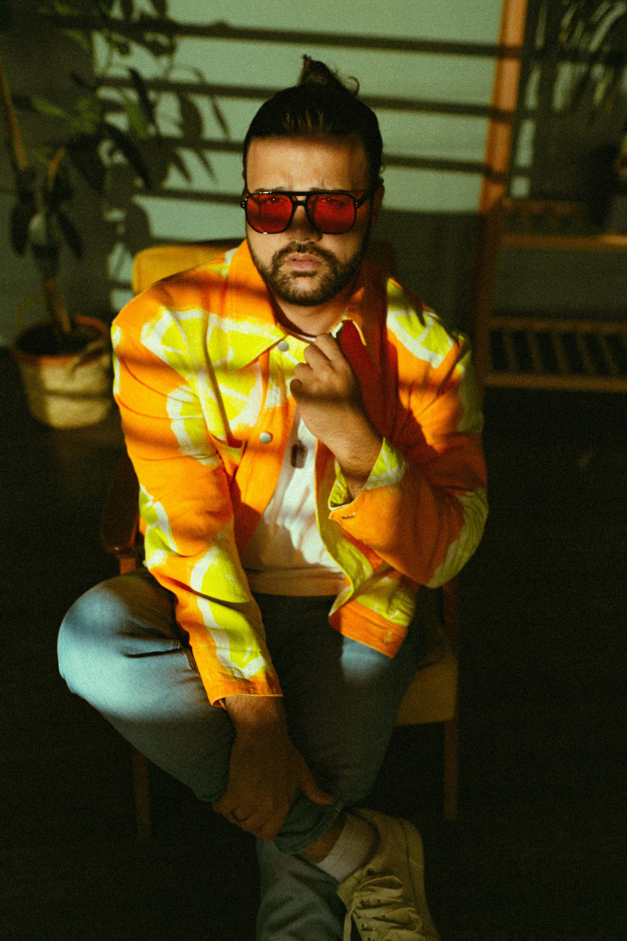 Fashionable man with sunglasses sits confidently in an orange and lemon-print jacket, embracing shadows and vintage vibes.