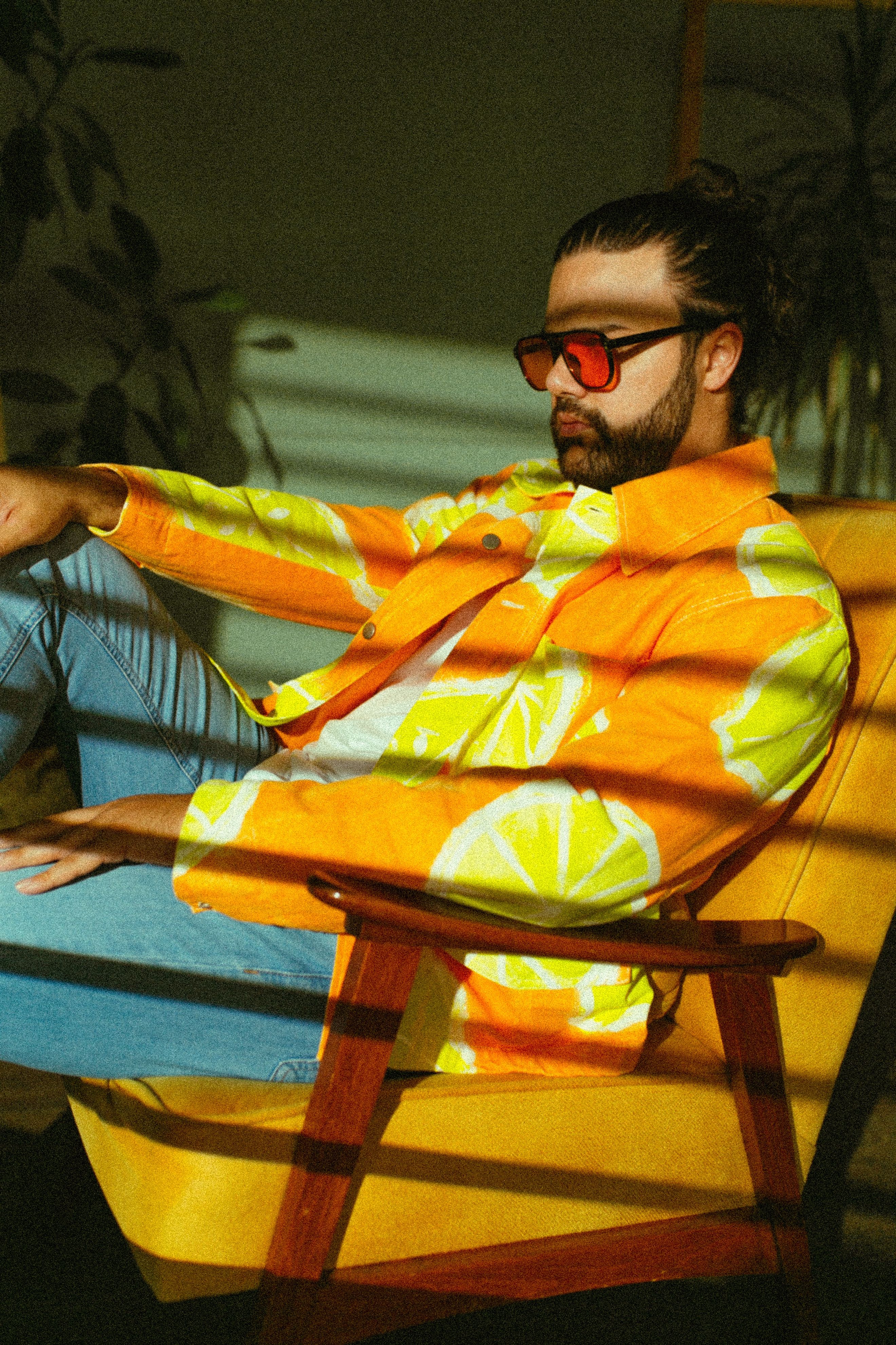 Stylish man in a vibrant orange shirt with sunglasses sitting in an armchair indoors.