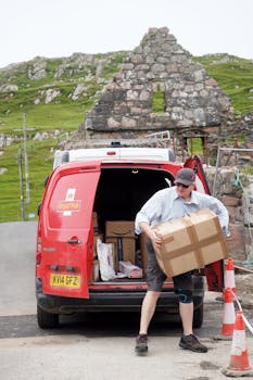 A postman delivers packages from a red Royal Mail van in a scenic rural location. 